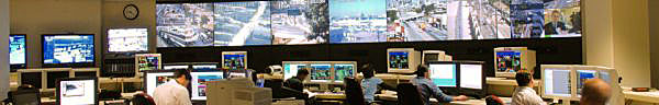 Photo shows a large room with about 10 work stations with people sitting in half of them.  Each has a computer monitor and keyboard, and is facing a wall that has 7 large screens showing different streets and communities.